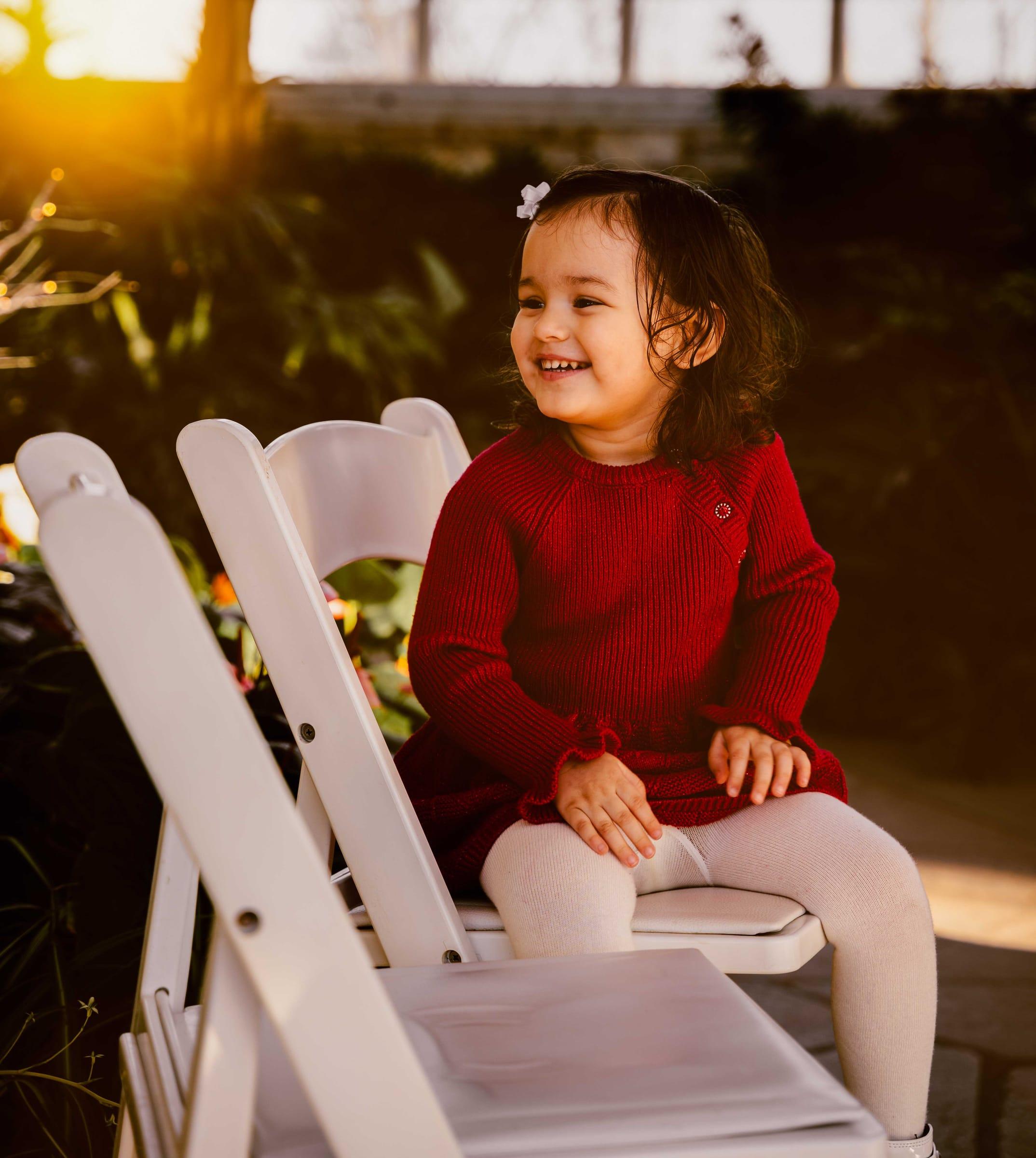 Toddler in a red dress smiles at golden hour on a white chair